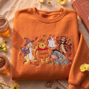 May include: An orange crewneck sweatshirt featuring an embroidered design of Winnie the Pooh characters dressed in Halloween costumes, including a pumpkin-clad Pooh, a witch Rabbit, a ghost Piglet, a vampire Tigger, and a mummy Eeyore.