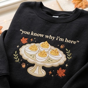 May include: A black crewneck sweatshirt featuring an embroidered design of four deviled eggs on a scalloped pedestal dish. The text "you know why i'm here" is embroidered above the dish, surrounded by small orange maple leaves, acorns, and stars.