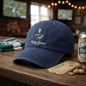 May include: A navy blue baseball cap featuring an embroidered graphic of a white goose wearing a green beanie and a green sweater with the word Philly on it. Below the goose, the text Philly Goose is embroidered in white script lettering.