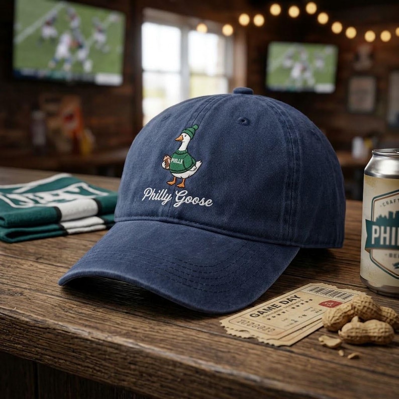 May include: A navy blue baseball cap featuring an embroidered graphic of a white goose wearing a green beanie and a green sweater with the word Philly on it. Below the goose, the text Philly Goose is embroidered in white script lettering.