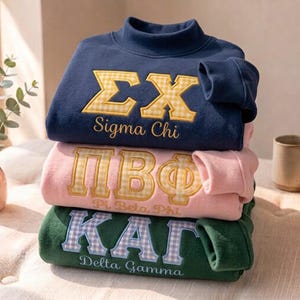Könnte beinhalten: Ein Stapel aus drei Sweatshirts mit Stehkragen in Marineblau, Hellrosa und Dunkelgrün. Jedes zeigt große, gestickte griechische Buchstaben mit Vichy-Muster und den Namen der jeweiligen Verbindung, der darunter in Schreibschrift gestickt ist.