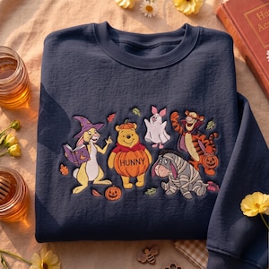 May include: A navy blue crewneck sweatshirt featuring an embroidered design of Winnie the Pooh characters dressed in Halloween costumes, including Rabbit as a wizard, Pooh as a pumpkin, Piglet as a ghost, Tigger as a vampire, and Eeyore as a mummy.