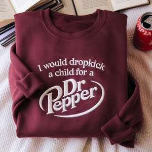 May include: Burgundy sweatshirt with the text "I would dropkick a child for a Dr Pepper" in white. A Dr Pepper can is visible in the upper right corner. The sweatshirt is folded on a white surface.