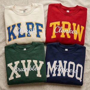 May include: Four crewneck sweatshirts in cream, red, dark green, and navy blue. Each features large, arched, block-letter appliqués in contrasting colors or patterns, with cursive white text embroidered over the center of the letters.