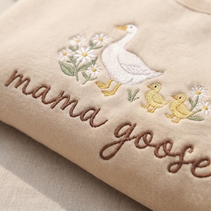 May include: A beige sweatshirt featuring embroidered details of a white goose, two yellow goslings, and white daisies with green leaves. The text "mama goose" is embroidered in brown cursive script across the front of the garment.