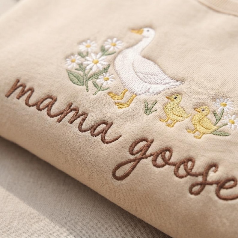 May include: A beige sweatshirt featuring embroidered details of a white goose, two yellow goslings, and white daisies with green leaves. The text "mama goose" is embroidered in brown cursive script across the front of the garment.