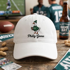 May include: A white baseball cap featuring a graphic of a goose wearing a green beanie and a green sweater with the word Philly printed on it. The goose is holding a brown football. The text Philly Goose is printed in black below the graphic.