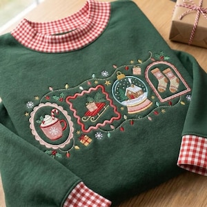 May include: A dark green sweatshirt featuring a red and white gingham collar and cuffs. The front displays embroidered holiday motifs including a mug of cocoa, a sled with a gift, a snow globe with a cabin, and two socks, all connected by string lights.