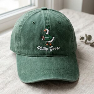 May include: A forest green baseball cap featuring an embroidered graphic of a white goose wearing a green beanie and a green sweater with the word Philly on it, while holding a brown football. The text Philly Goose is embroidered below.