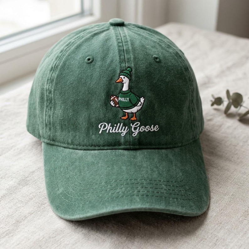 May include: A forest green baseball cap featuring an embroidered graphic of a white goose wearing a green beanie and a green sweater with the word Philly on it, while holding a brown football. The text Philly Goose is embroidered below.
