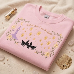 Op de afbeelding: Een lichtroze sweater met ronde hals en een borduursel van twee figuren in een boot op het water, omringd door zwevende lantaarns, een paarse toren en gele bloemen. De tekst "And at last I see the light" is in goud geborduurd.