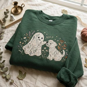 May include: A dark green crewneck sweatshirt featuring an embroidered design of a white lace ghost and a white lace dog surrounded by small flowers and stars. The text BOO-P is embroidered in white above the dog, with a small star accent.