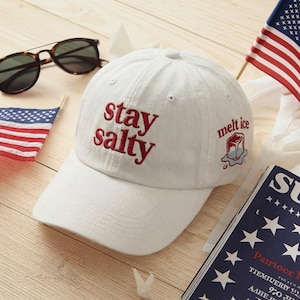 Può includere: Cappellino da baseball bianco con la scritta ricamata "stay salty" in rosso. Il cappellino presenta anche una piccola grafica di ghiaccio che si scioglie e le parole "melt ice". Occhiali da sole e una piccola bandiera americana sono visibili.