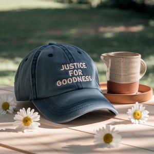 May include: A blue baseball cap with the text "JUSTICE FOR GOODNESS" embroidered in white. The cap is on a wooden surface with a ceramic pitcher and daisies. The background is blurred green.