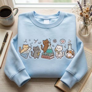 May include: Pale blue sweatshirt with a design of cartoon cats in a chemistry lab. The cats are shown with beakers, test tubes, and lab coats. The words "LAB NOTES CHEMISTRY" are printed on a book. The sweatshirt has a crew neck and long sleeves.