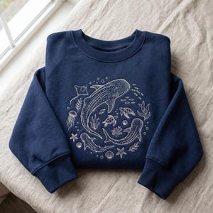 May include: A navy blue crewneck sweatshirt featuring a white line art graphic of marine life, including whale sharks, sea turtles, a stingray, a jellyfish, starfish, seashells, and seaweed arranged in a circular pattern on the chest.