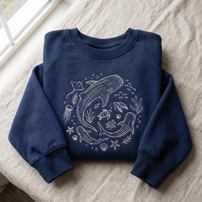 May include: A navy blue crewneck sweatshirt featuring a white line art graphic of marine life, including whale sharks, sea turtles, a stingray, a jellyfish, starfish, seashells, and seaweed arranged in a circular pattern on the chest.