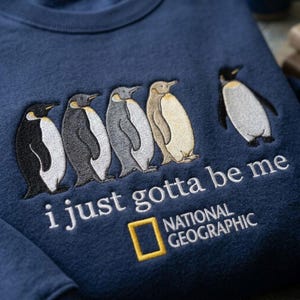 May include: A navy blue sweatshirt featuring an embroidered design of five penguins in a row. Below the penguins, the white text reads "i just gotta be me" above the yellow National Geographic logo and white text.