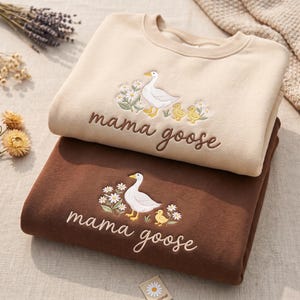 May include: Two crewneck sweatshirts, one beige and one brown, each featuring an embroidered design of a white goose with yellow goslings and white daisies. The text "mama goose" is embroidered in brown script below the goose illustration.