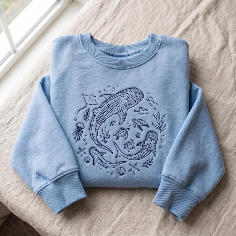 May include: A light blue crewneck sweatshirt featuring a dark blue embroidered design of marine life, including whale sharks, sea turtles, a jellyfish, a stingray, starfish, seashells, and seaweed arranged in a circular pattern on the front.