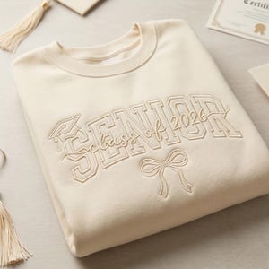 Könnte beinhalten: Cremefarbenes Sweatshirt mit dem gestickten Text "SENIOR Class of 2026" in einer College-Schriftart, zusammen mit einem Abschlusskappen- und Schleifendesign. Das Sweatshirt ist ordentlich gefaltet, mit Quasten und einer Urkunde im Hintergrund.