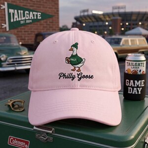 May include: A light pink baseball cap featuring an embroidered graphic of a white goose wearing a green beanie and a green sweater with the word Philly on it, while holding a brown football. The text Philly Goose is embroidered below the goose.