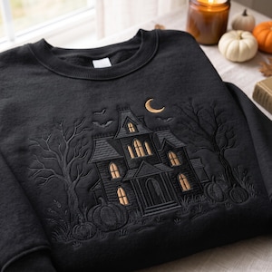 Pode incluir: Uma camisola preta de gola redonda com bordado detalhado de uma casa assombrada, árvores sem folhas e abóboras. O design inclui uma lua crescente e morcegos, com fio amarelo-dourado a destacar as janelas da casa.