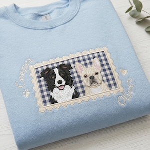 Pode incluir: Uma camisola azul clara de gola redonda com um design bordado de selo postal sobre um fundo axadrezado azul-marinho e branco. O bordado apresenta um Border Collie preto e branco e um Bulldog francês creme, com os nomes Cooper e Oliver.