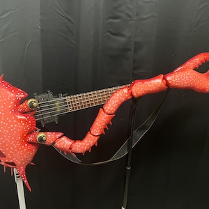 May include: A unique, custom-made electric bass guitar shaped like a red lobster. The instrument features a detailed lobster claw, body, and tail design, with a black neck and strings. The lobster's body has a textured, spotted pattern.