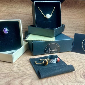 May include: Jewelry display featuring rings, a necklace, and boxes. A silver ring with a purple stone sits in a box. A gold necklace with a white stone is in a black box. Other rings with gemstones are also displayed.
