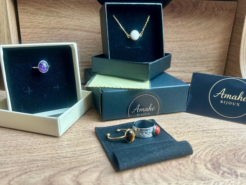 May include: Jewelry display featuring rings, a necklace, and boxes. A silver ring with a purple stone sits in a box. A gold necklace with a white stone is in a black box. Other rings with gemstones are also displayed.