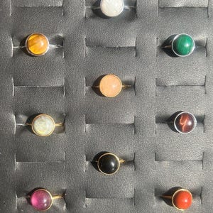 May include: A collection of gemstone rings displayed in a black tray. The rings feature various colored stones, including white, yellow, green, pink, and black, set in gold-colored bands. The stones are round and polished.