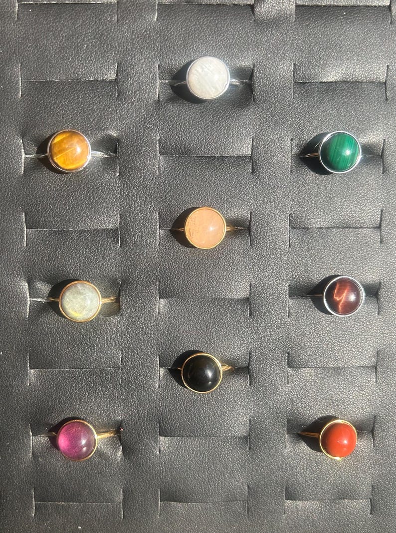 May include: A collection of gemstone rings displayed in a black tray. The rings feature various colored stones, including white, yellow, green, pink, and black, set in gold-colored bands. The stones are round and polished.
