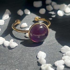 May include: A gold-colored adjustable ring featuring a large, round, deep purple gemstone. The ring's band is open, with small gold spheres at the ends. The gemstone is set in a gold bezel, creating a striking contrast. The ring is displayed on a dark gray surface.