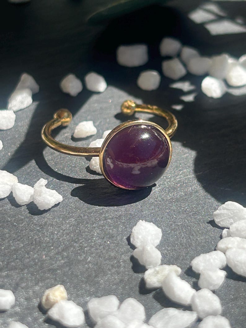 May include: A gold-colored adjustable ring featuring a large, round, deep purple gemstone. The ring's band is open, with small gold spheres at the ends. The gemstone is set in a gold bezel, creating a striking contrast. The ring is displayed on a dark gray surface.