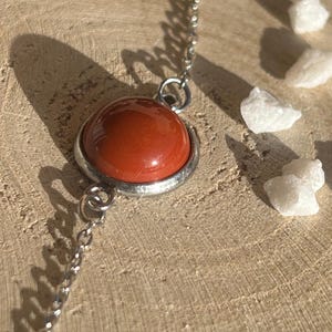May include: A close-up of a necklace featuring a large, round, reddish-orange gemstone pendant set in a silver-colored metal frame. The pendant hangs from a delicate silver chain. White, irregularly shaped stones are scattered around the necklace on a wooden surface.