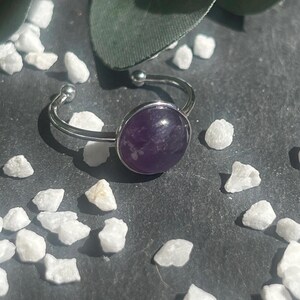 May include: A silver-toned adjustable ring featuring a large, oval, deep purple amethyst stone. The ring is set against a dark gray surface, surrounded by small white stones and a green leaf.