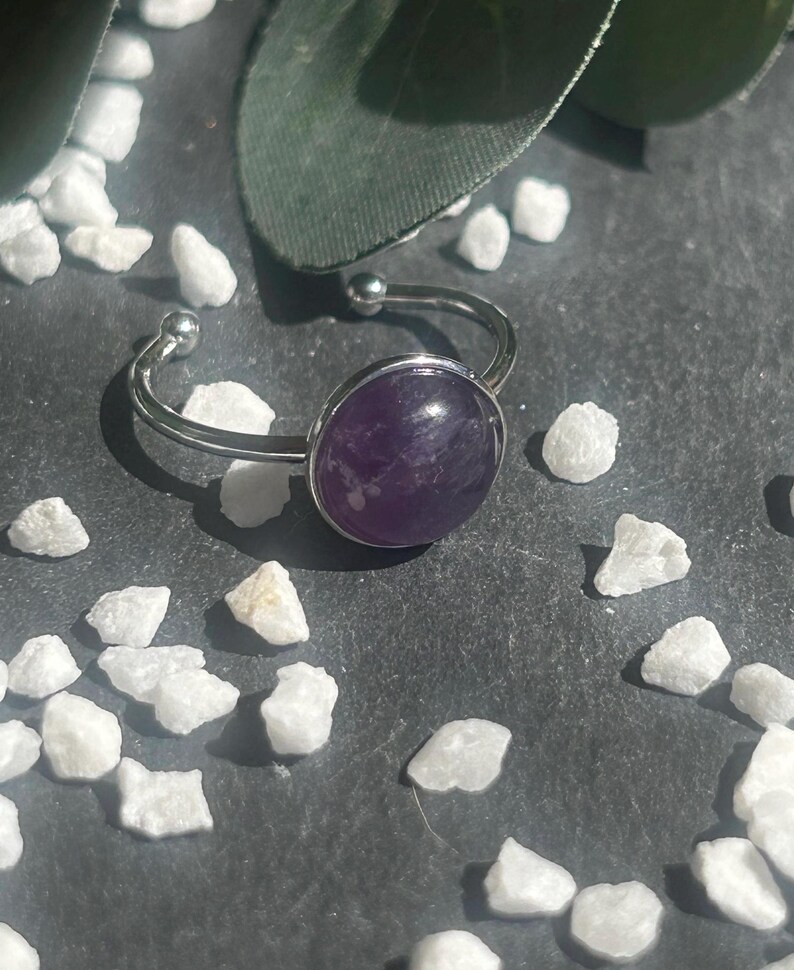 May include: A silver-toned adjustable ring featuring a large, oval, deep purple amethyst stone. The ring is set against a dark gray surface, surrounded by small white stones and a green leaf.