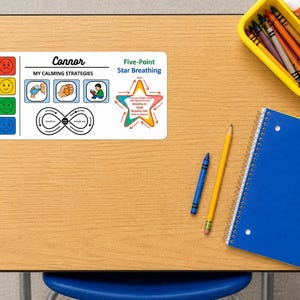 May include: A desk with school supplies, including a blue notebook, pencils, crayons, and a yellow tray. A white card with the name "Connor" and calming strategies is also on the desk. The card features illustrations and text about managing emotions.