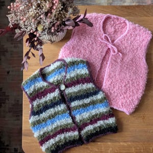 May include: Two handmade knitted vests. One is pink with a tie closure, the other is striped with blue, green, and burgundy. A single button secures the striped vest. The vests are displayed on a wooden surface.
