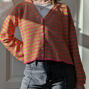 May include: A long-sleeved V-neck cardigan featuring horizontal stripes in bright pink and lime green. The knit garment has a button-down front with six small pink buttons, a ribbed hem, and ribbed cuffs, worn over a white ribbed undershirt.