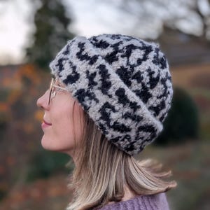 Könnte beinhalten: Eine gestrickte Beanie-Mütze mit einem schwarz-weißen Muster. Die Mütze wird von einer Person mit blondem Haar und Brille getragen. Die Person trägt einen lila Pullover. Der Hintergrund ist verschwommen mit Herbstfarben.