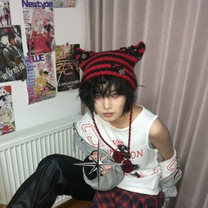 May include: A red and black striped cat ear hat with metal accents. The person is wearing a white tank top with the text "VAS ON" and a red and black plaid skirt. Anime posters are visible in the background.