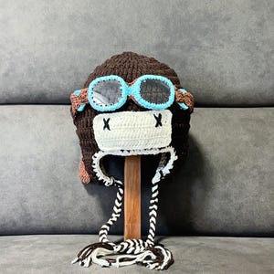 May include: A brown crocheted hat with a white and blue aviator goggles design. The hat features a white face with black 'X' markings and brown and white braided ties. The hat is displayed on a wooden stand.