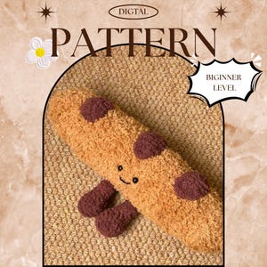 Puede incluir: Un cojín afelpado en forma de galleta, marrón y granate, con una cara sonriente y dos pies granates. La imagen incluye las palabras "DIGITAL PATTERN" y "BEGINNER LEVEL" en un marco decorativo.