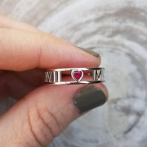 May include: A silver ring with Roman numerals and a red heart-shaped gemstone in the center.