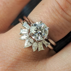 May include: A close-up of a silver ring with a large round diamond centre stone and smaller diamonds set in a delicate, floral design. The ring is worn on a person's finger.