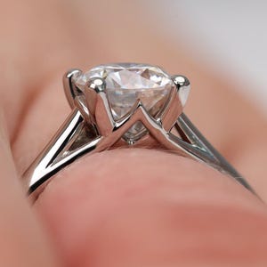 May include: Close-up of a silver engagement ring with a large, clear, round-cut diamond. The ring's band is thin and polished, with a four-prong setting holding the diamond securely. The background is a soft, blurred peach color.