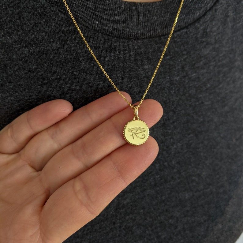 May include: Gold necklace with a round pendant featuring the Eye of Horus symbol.
