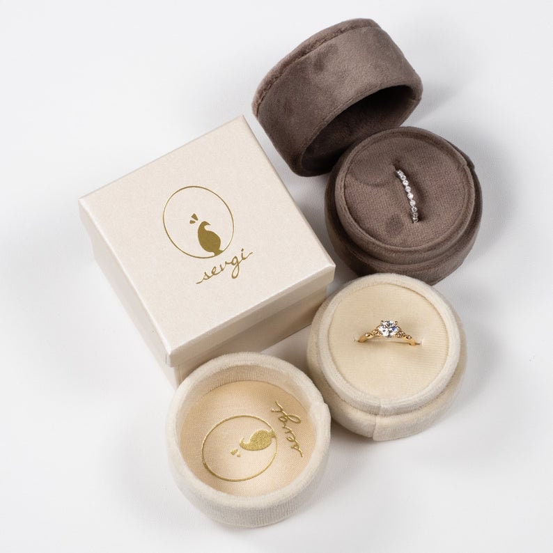 May include: Two ring boxes, one brown velvet and one cream colored, with a gold ring inside each. The cream colored box has the text "sergi" on the side.
